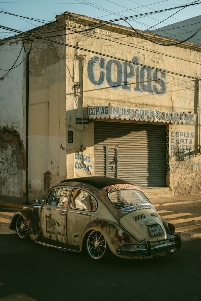 A rusty vintage car parked in front of an old building with faded signage, under evening light.